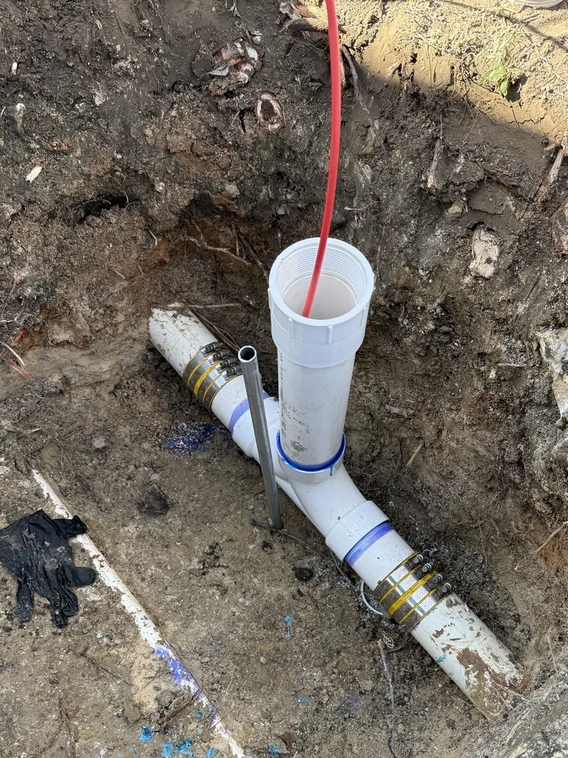 A newly installed white PVC sewer line in a trench with a cleanout riser fitted and a line fed down into it.
